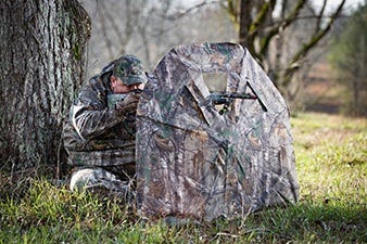 Hunter Sitting Behind Ground Blind With Rifle Raised to Shoot 